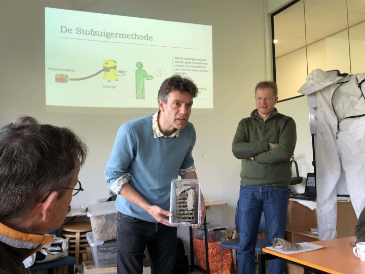 Een man met een kunststof bakje in zijn handen, met daarin de raten en een deel van het omhulsel van een wespennest. De raten zijn als etages boven elkaar gebouwd. De man staat duidelijk gepassioneerd te vertellen. Op de achtergrond staat een man toe te kijken, en zie je een slide uit een PowerPoint met als titel 'De stofzuigermethode'. Rechts zie je op de achtergrond nog een wit pak hangen, dat is een imkeroverall.