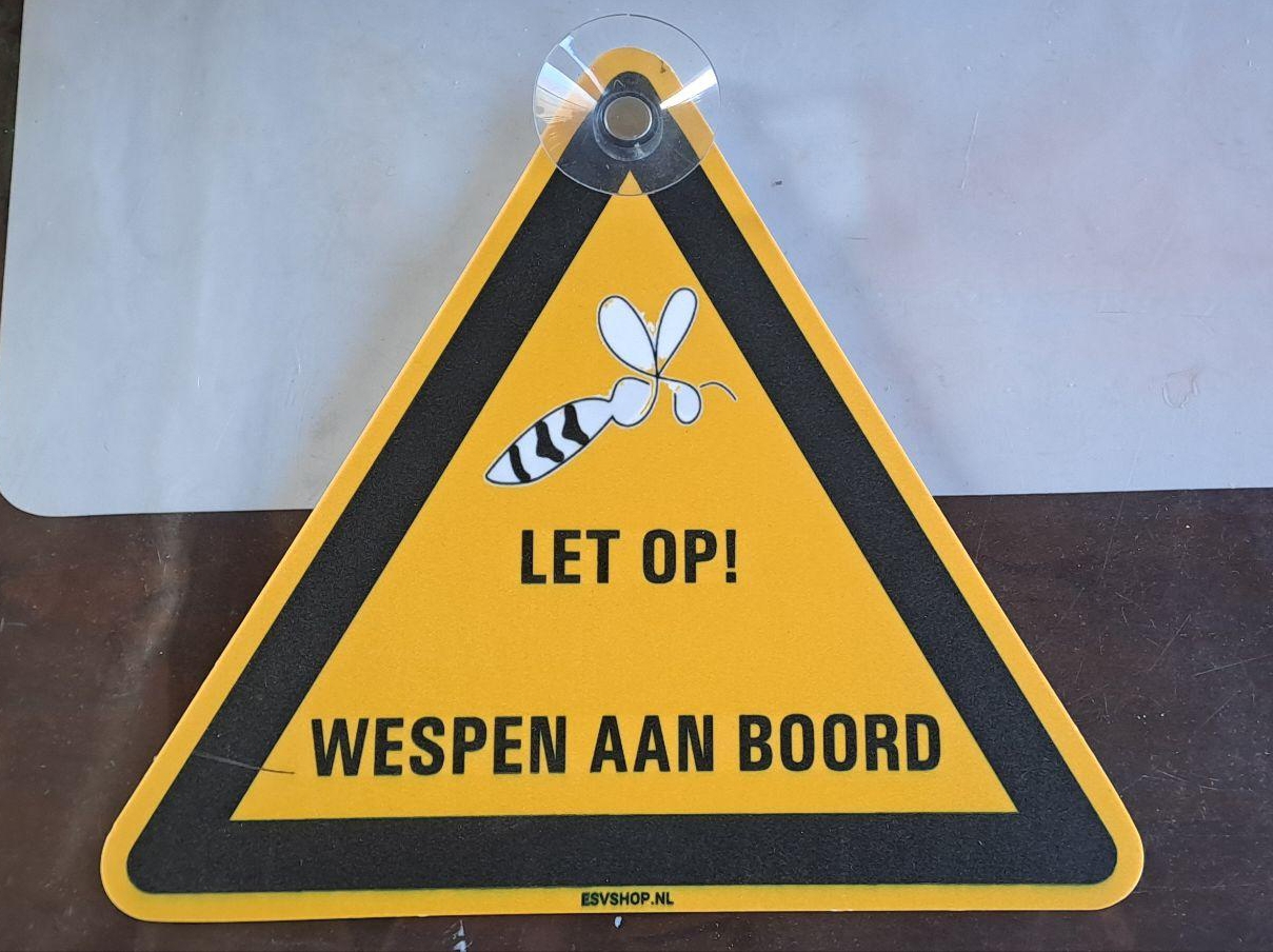 Gele driehoek met daarin een zwarte driehoek waarin een gestyleerde wesp en daaronder in hoofdletters de tekst "Let op! Wespen aan boord" staan. In de bovenste punt zit een zuignap bevestigd.