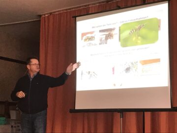 Scherm met dia van een presentatie met daarop enkele foto's van wespen en de tekst 'Wie siehen die Tiere aus?' Links van het scherm staat een man die duidelijk aan het vertellen is over deze dia, met zijn linkerhand die wijst op het scherm.