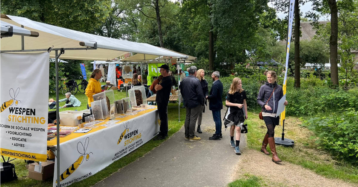 Kraam met wit zeil op het dak en over de tafel een doek met het logo van de Wespenstichting aan de voorkant. Op de kraam staan enkele wespennesten, achter de kraam staat een vrouw met een gele hoodie aan met iemand aan de andere kant van de kraam te praten. Daarachter staat nog een groepje van drie mensen te plaatsen en er lopen twee dames voorbij. Op de achtergrond zie je groen van bomen en planten.
